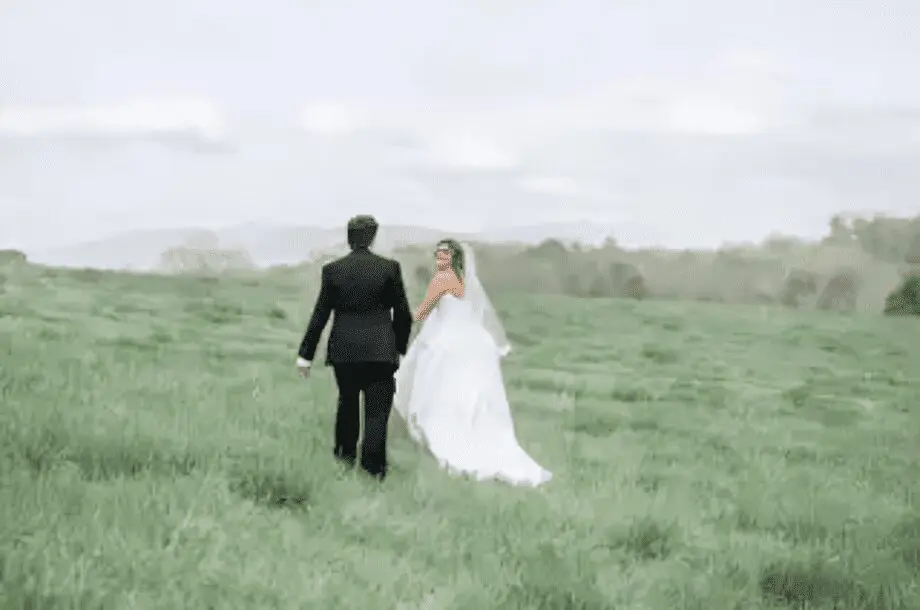 Bride and groom walking through grassy field.